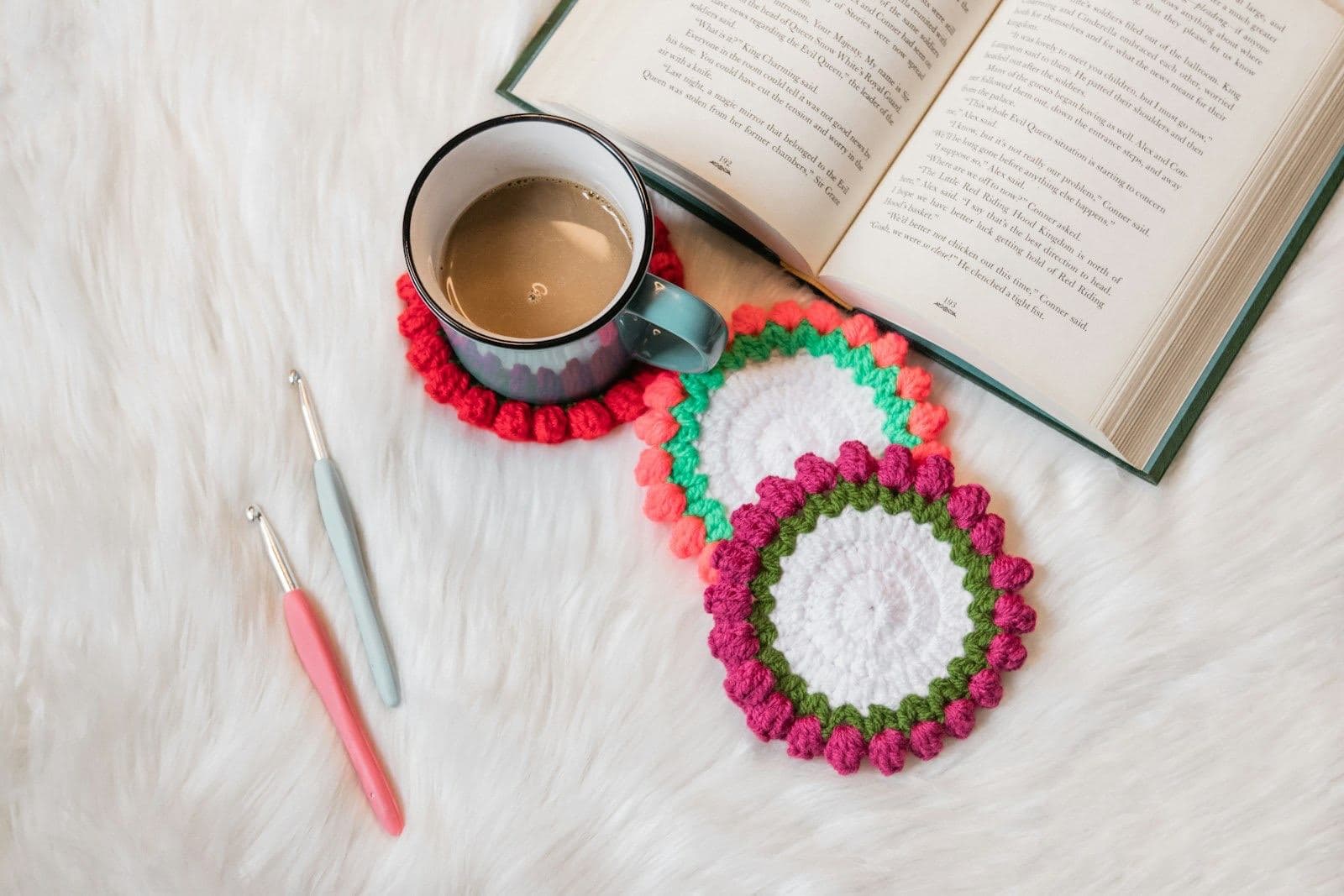 Round crochet coasters with bobble edges beside a mug of coffee, two crochet hooks, and an open book