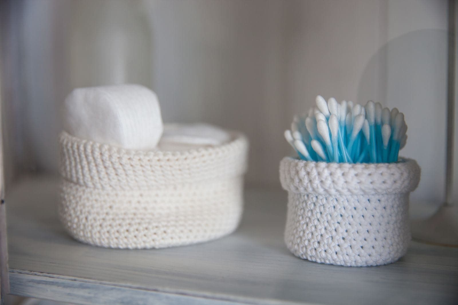Two small cream crochet baskets on a white shelf, holding cotton pads and cotton buds