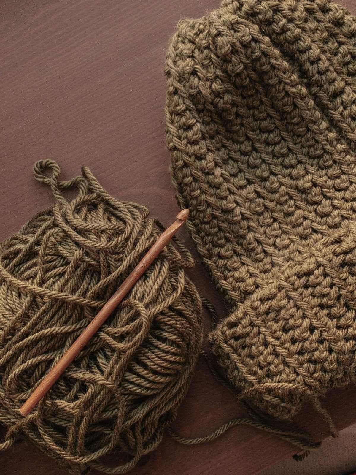 An olive green crochet beanie with a folded brim beside a skein of matching yarn and a wooden crochet hook