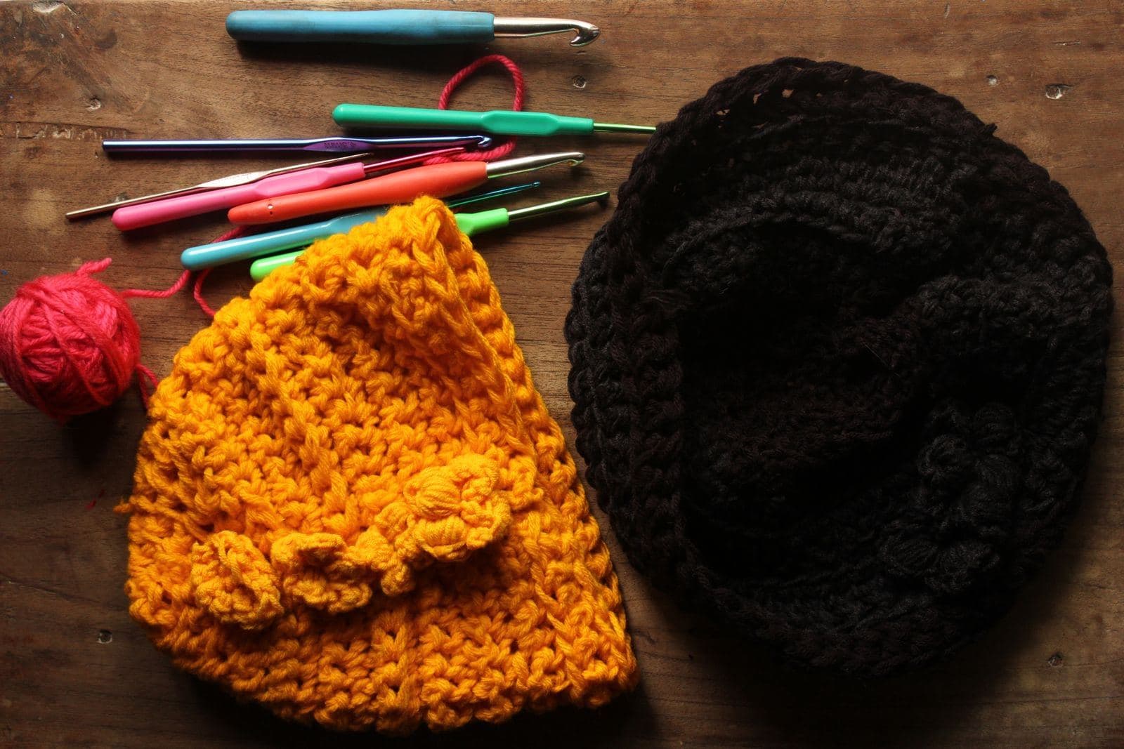 A mustard crochet beanie with flower details and a black crochet hat beside colorful crochet hooks