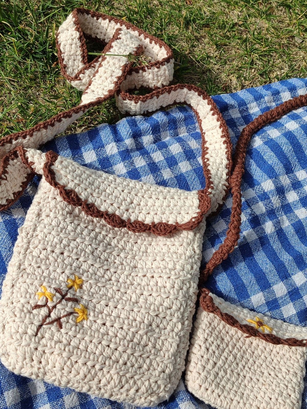 A cream crochet shoulder bag with brown trim and small embroidered flowers on a blue gingham picnic blanket