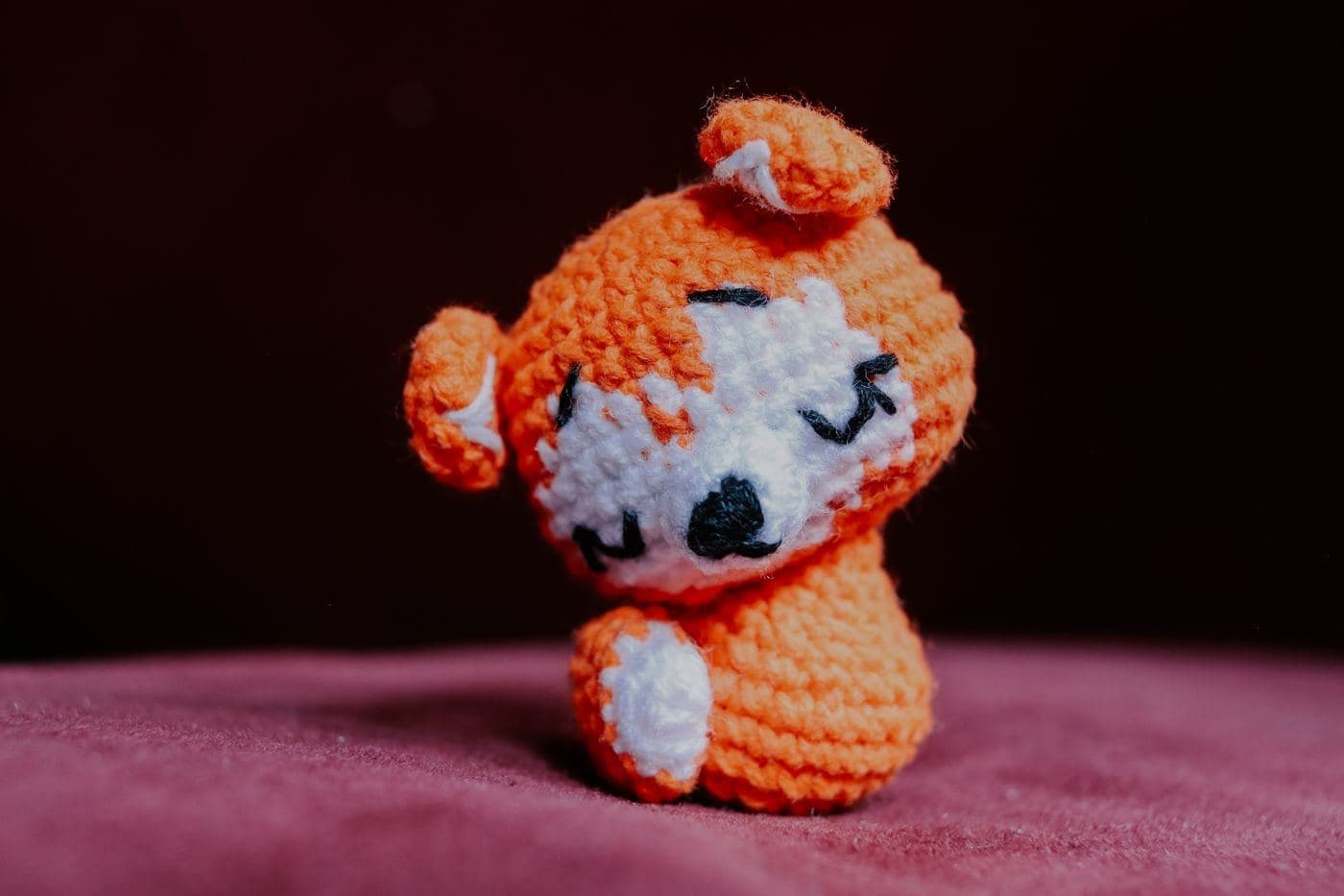 A small orange and white amigurumi animal with a round crocheted head on a pink cushion