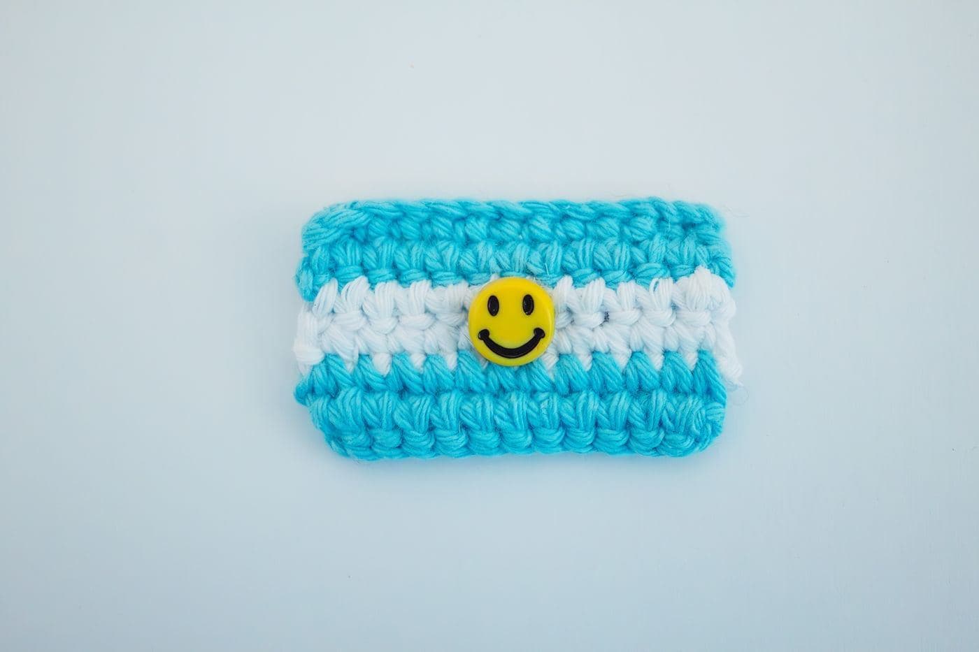 A small blue and white striped crochet pouch with a yellow smiley-face button on a pale blue background
