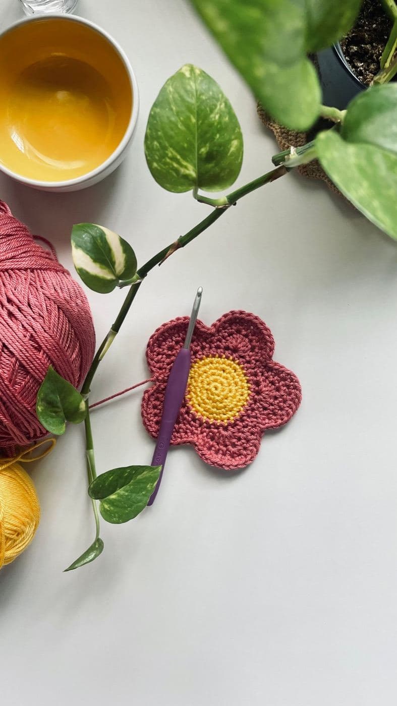 A red crochet flower with five rounded petals and a yellow center beside a purple crochet hook, pink yarn, and a plant