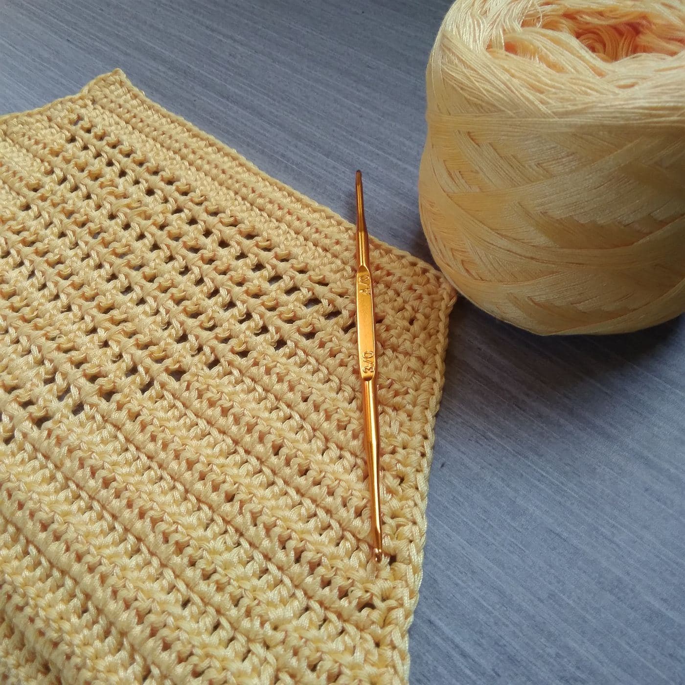Rows of pale yellow crochet fabric with a gold crochet hook and a ball of matching yarn