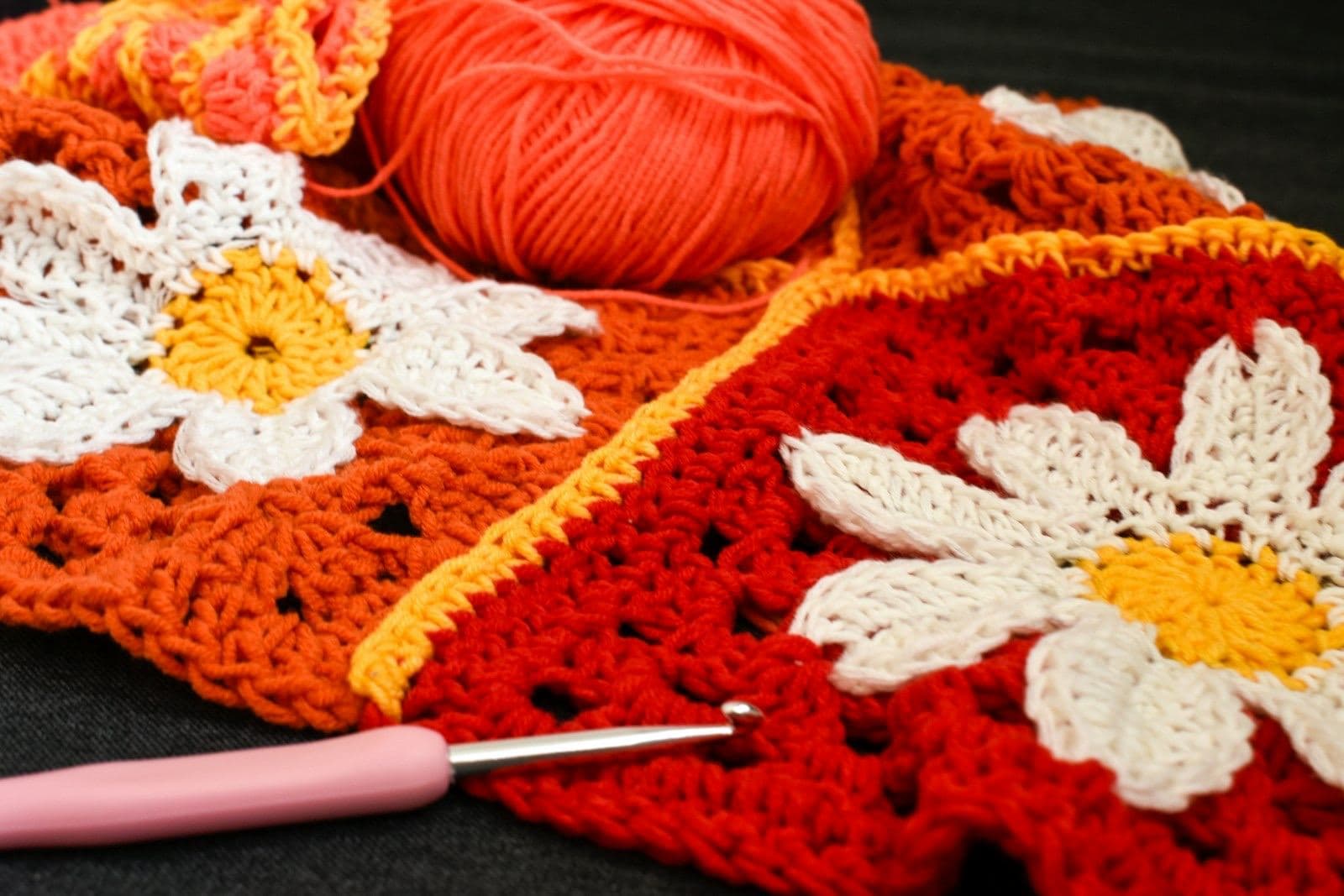 Red and orange crochet squares with white daisy motifs, a ball of yarn, and a pink crochet hook