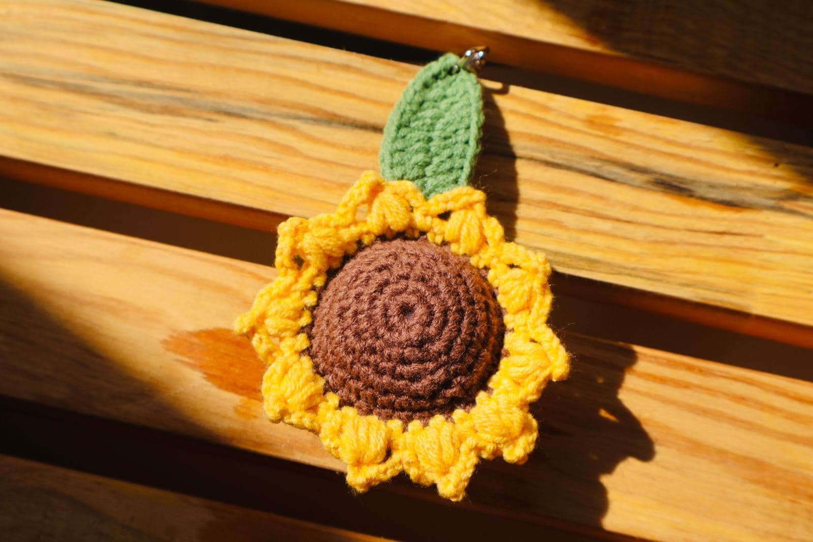A crochet sunflower keychain with a brown center, yellow petals, and a green leaf on a sunlit wooden bench